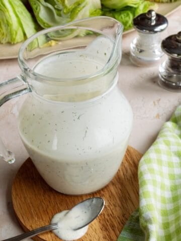 Pitcher of copycat Chili's ranch recipe on a wooden coaster with a platter of iceberg lettuce wedges in the background.