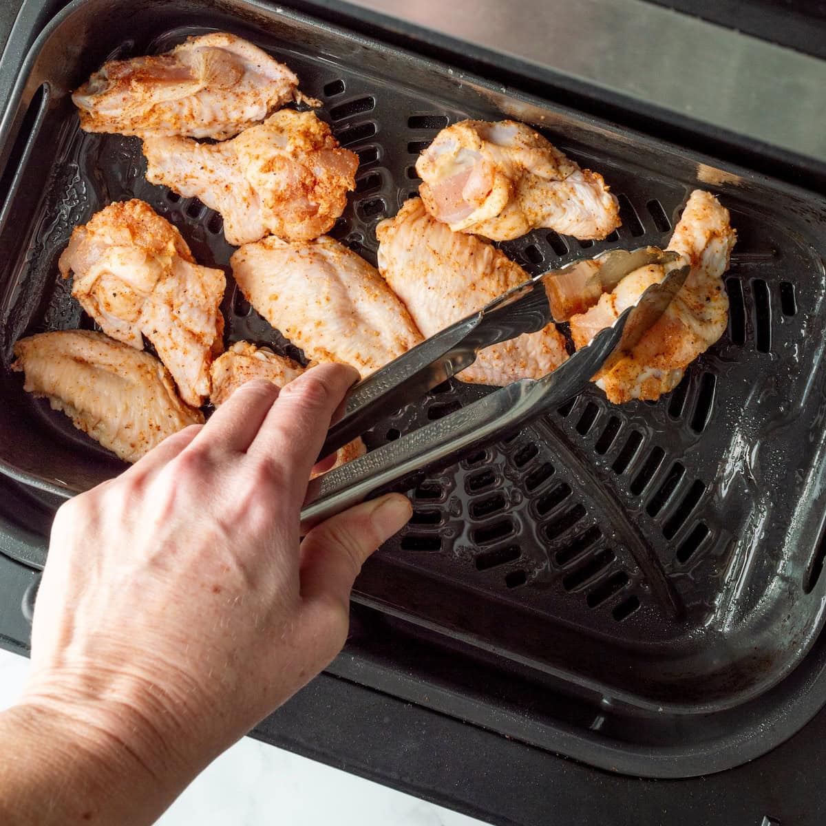 Adding seasoned chicken wings to an air fryer with metal tongs.