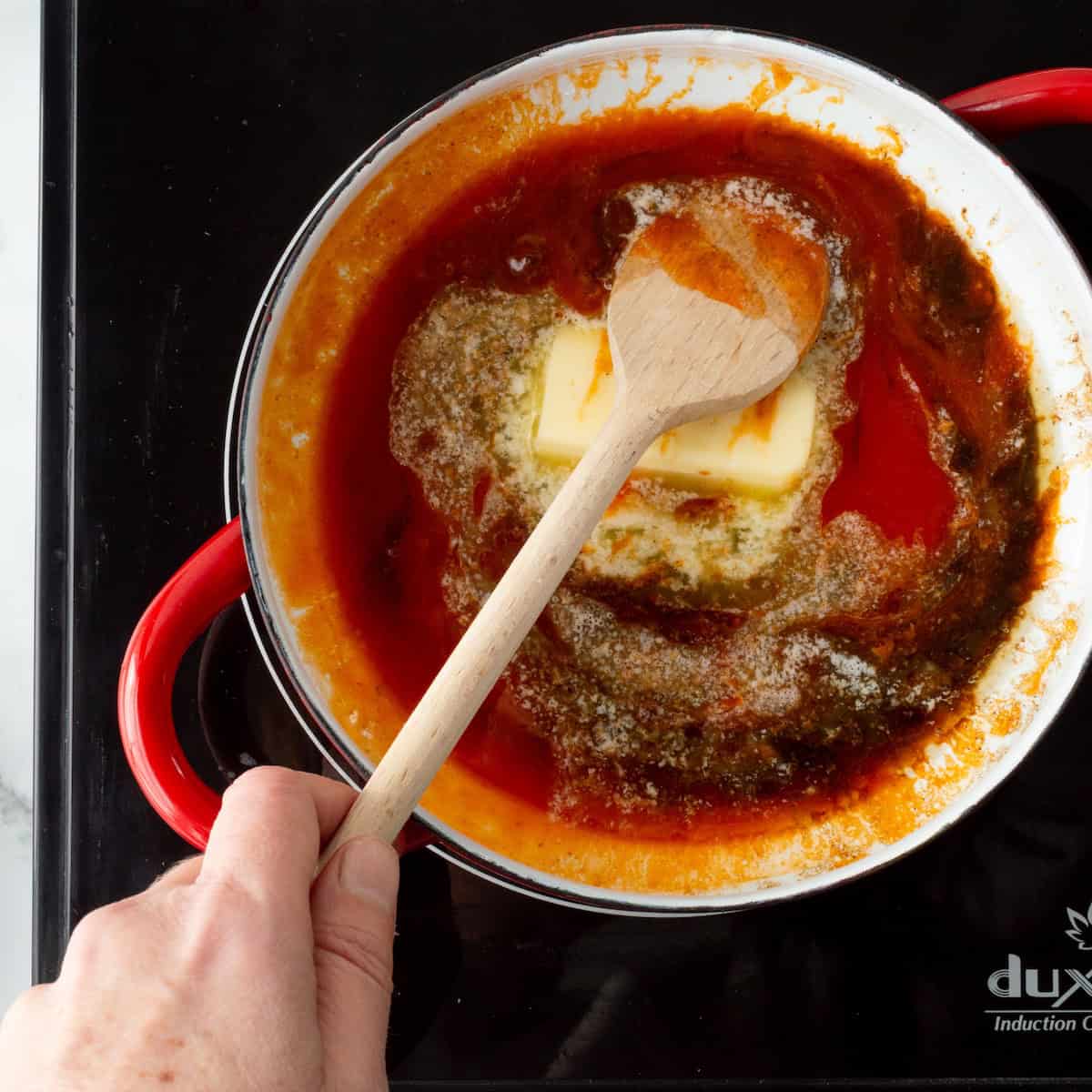Stirring together butter, buffalo hot wing sauce, and Worcestershire sauce in a red handled saucepan.