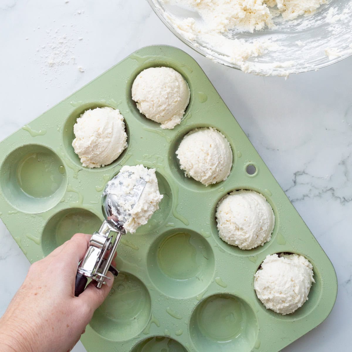 Scooping 3 ingredient biscuit dough into a green muffin pan with an ice cream scooper.