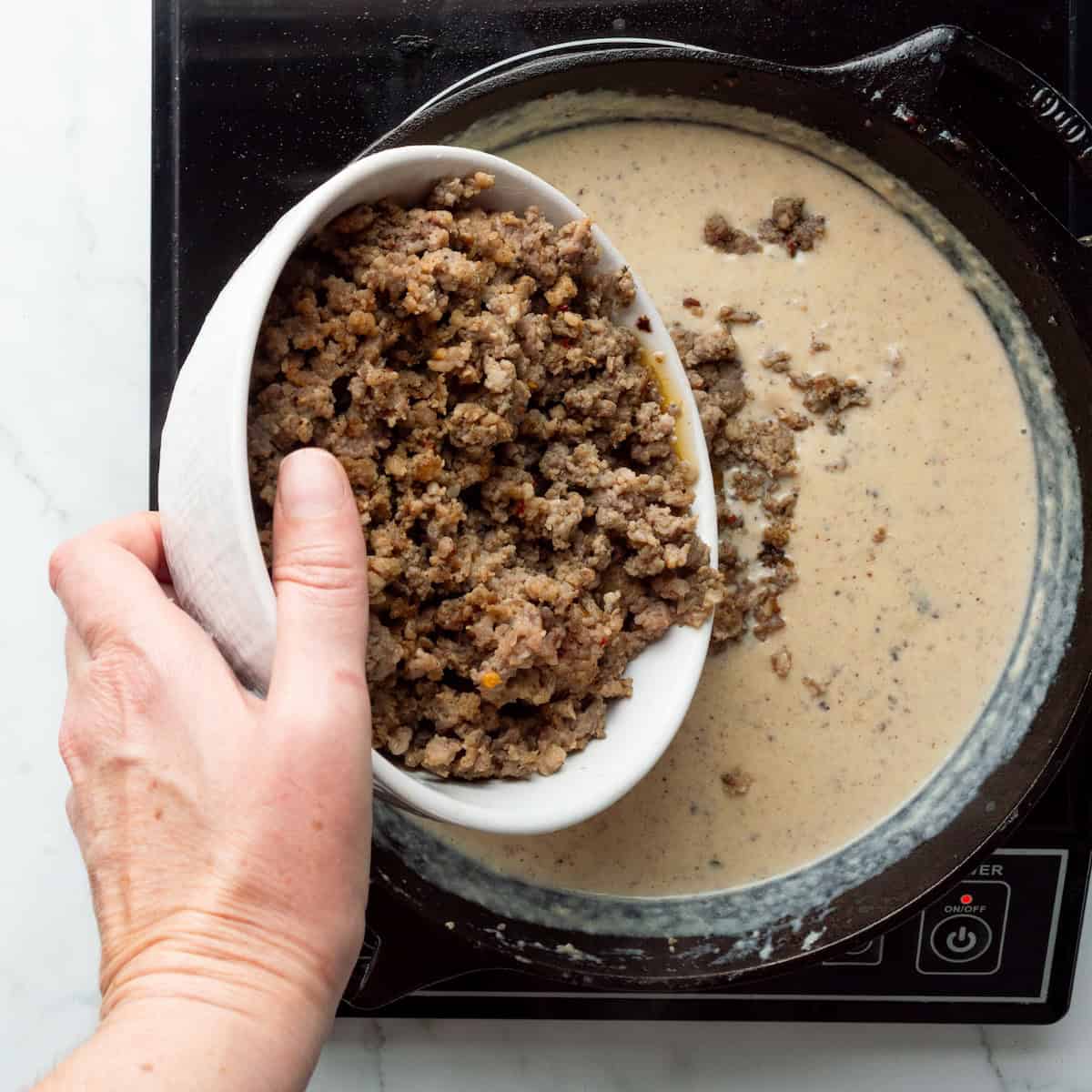 Adding browned breakfast sausage to a cast iron skillet of gravy.