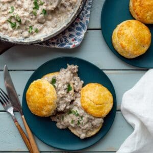 A skillet of homestyle sausage gravy with two blue plates, one topped with biscuits and gravy and another with just biscuits.