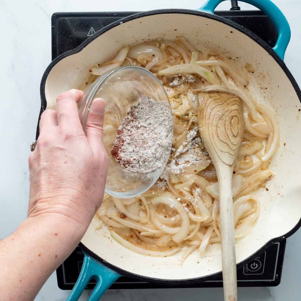 Adding seasoned flour to cooked onions in a blue enamel cast iron skillet.
