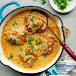 Southern smothered chicken thighs in a blue handled skillet on a woven trivet with a plate of parsley to the side.