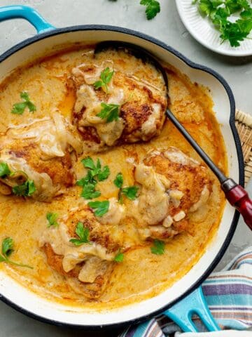 Southern smothered chicken thighs in a blue handled skillet on a woven trivet with a plate of parsley to the side.