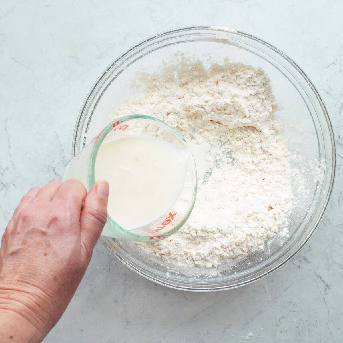 Pouring buttermilk into a glass bowl of flour and butter mixture for biscuits.