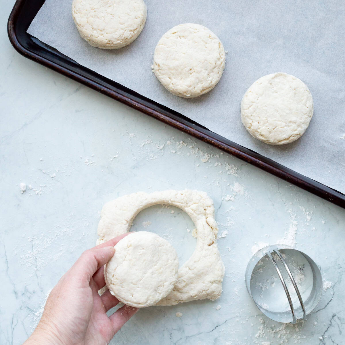 Using a biscuit cutter to cut biscuits out of a small square of dough and place them on a parchment lined baking sheet.