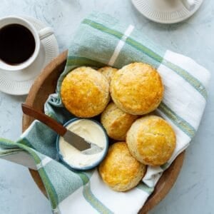 A wooden bowl of copycat Popeye's buttermilk biscuits with a small blue bowl of butter along with cups of coffee.