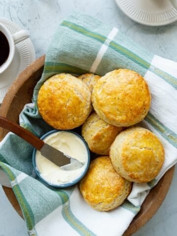 A wooden bowl of copycat Popeye's buttermilk biscuits with a small blue bowl of butter along with cups of coffee.