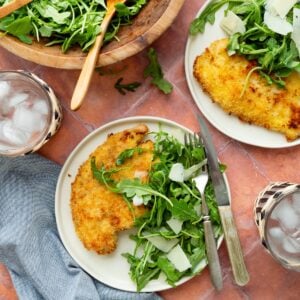 Breaded chicken cutlets on white plates topped with arugula salad and shaved Parmesan with a wooden bowl of arugula salad to the side.