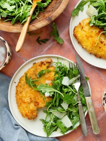 Breaded chicken cutlets on white plates topped with arugula salad and shaved Parmesan with a wooden bowl of arugula salad to the side.