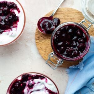 Homemade blueberry compote in a glass jar on a round wooden board surrounded by bowls of Greek yogurt topped with compote and a blue cloth.