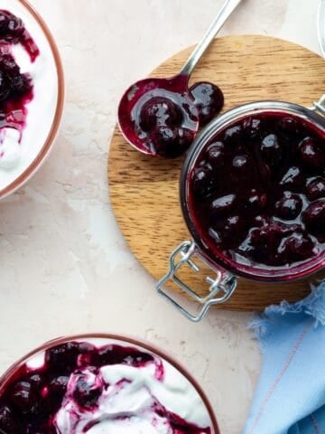Homemade blueberry compote in a glass jar on a round wooden board surrounded by bowls of Greek yogurt topped with compote and a blue cloth.