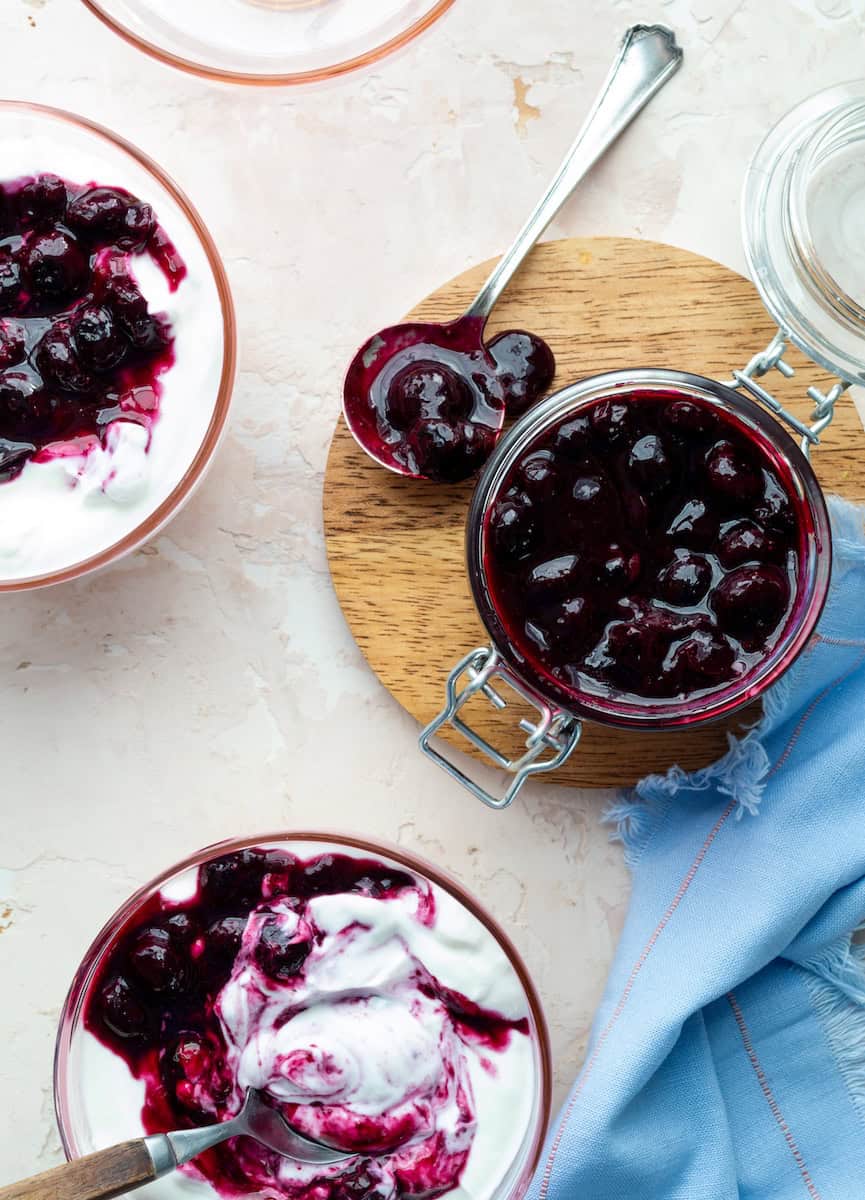 Blueberry compote in a jam jar on a round wooden board surrounded by bowls of Greek yogurt topped with compote.