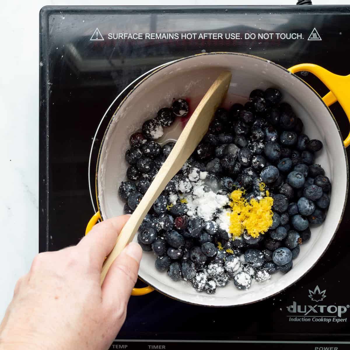 Cooking blueberries with sugar, lemon zest, cornstarch, lemon juice, and water in a yellow Dansk saucepan.