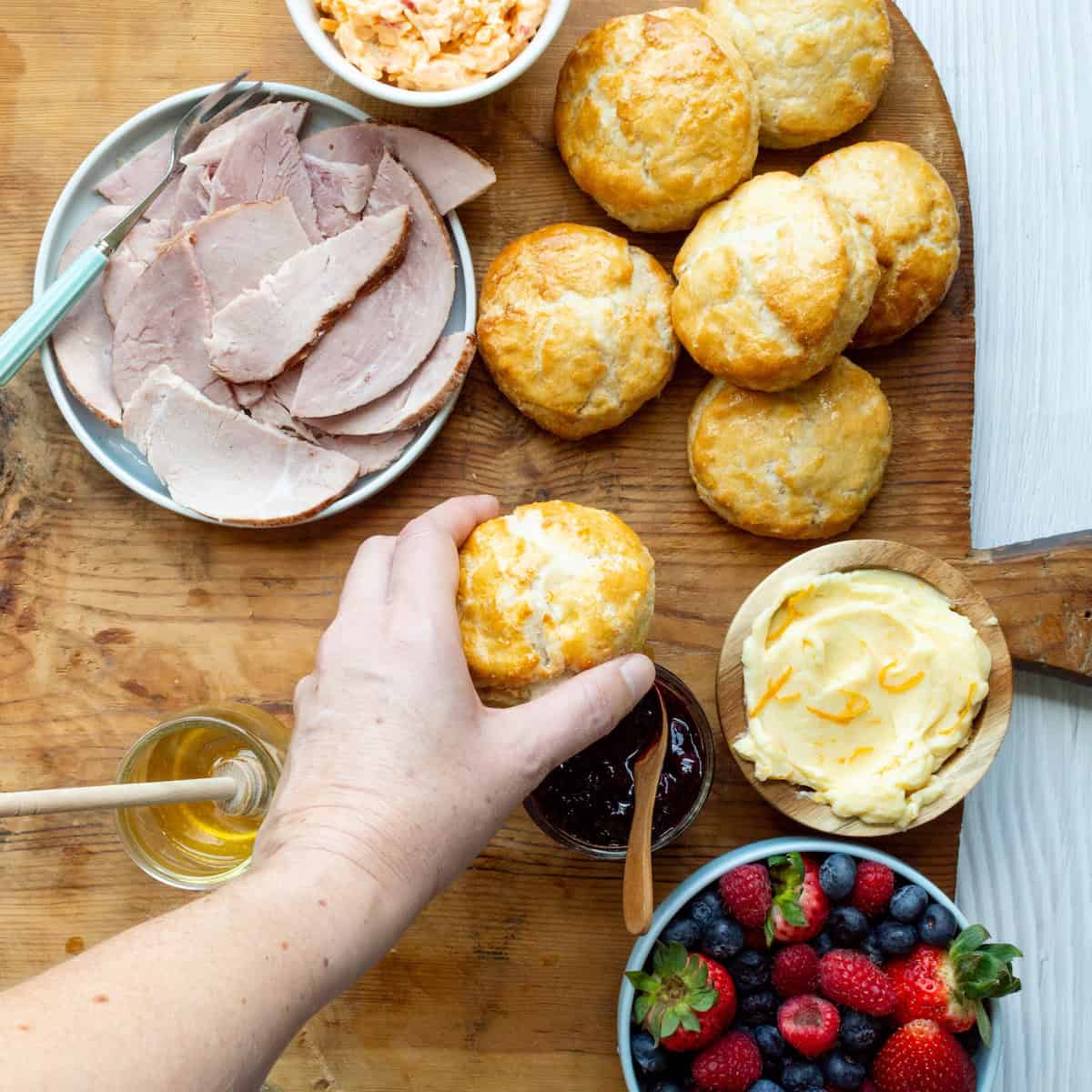 Adding buttermilk biscuits to a wooden charcuterie board with ham, pimento cheese, honey butter, blueberry compote, honey, and fresh blueberries, strawberries, and raspberries.