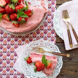 Strawberry cake with strawberry filling and strawberry buttercream on a white platter with a slice on white scalloped plate.