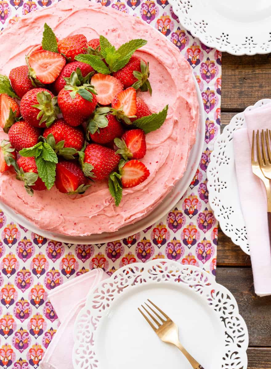 A strawberry and mint topped strawberry cake on a white platter with white plates on the side.