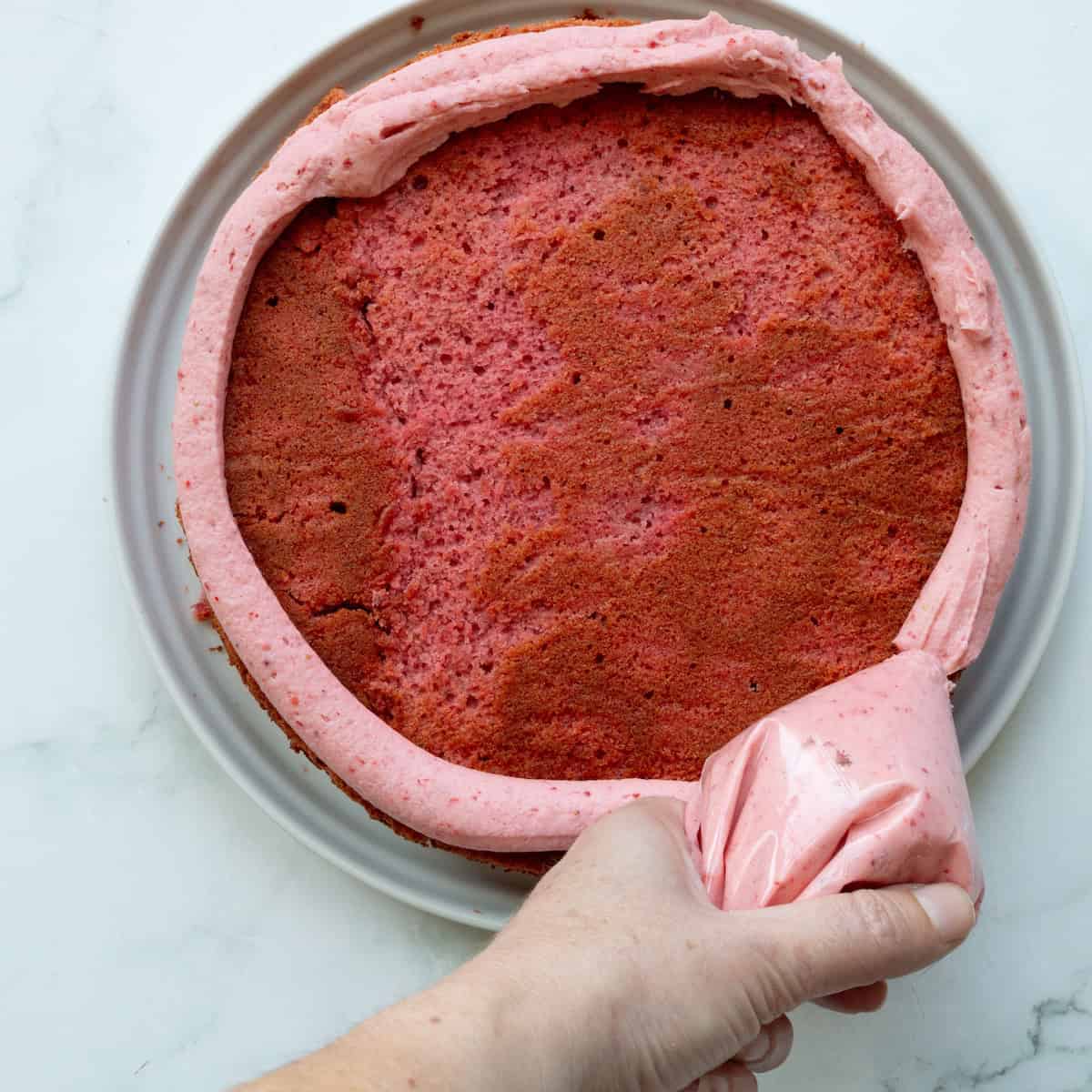 Piping strawberry buttercream around the edge of strawberry cake.