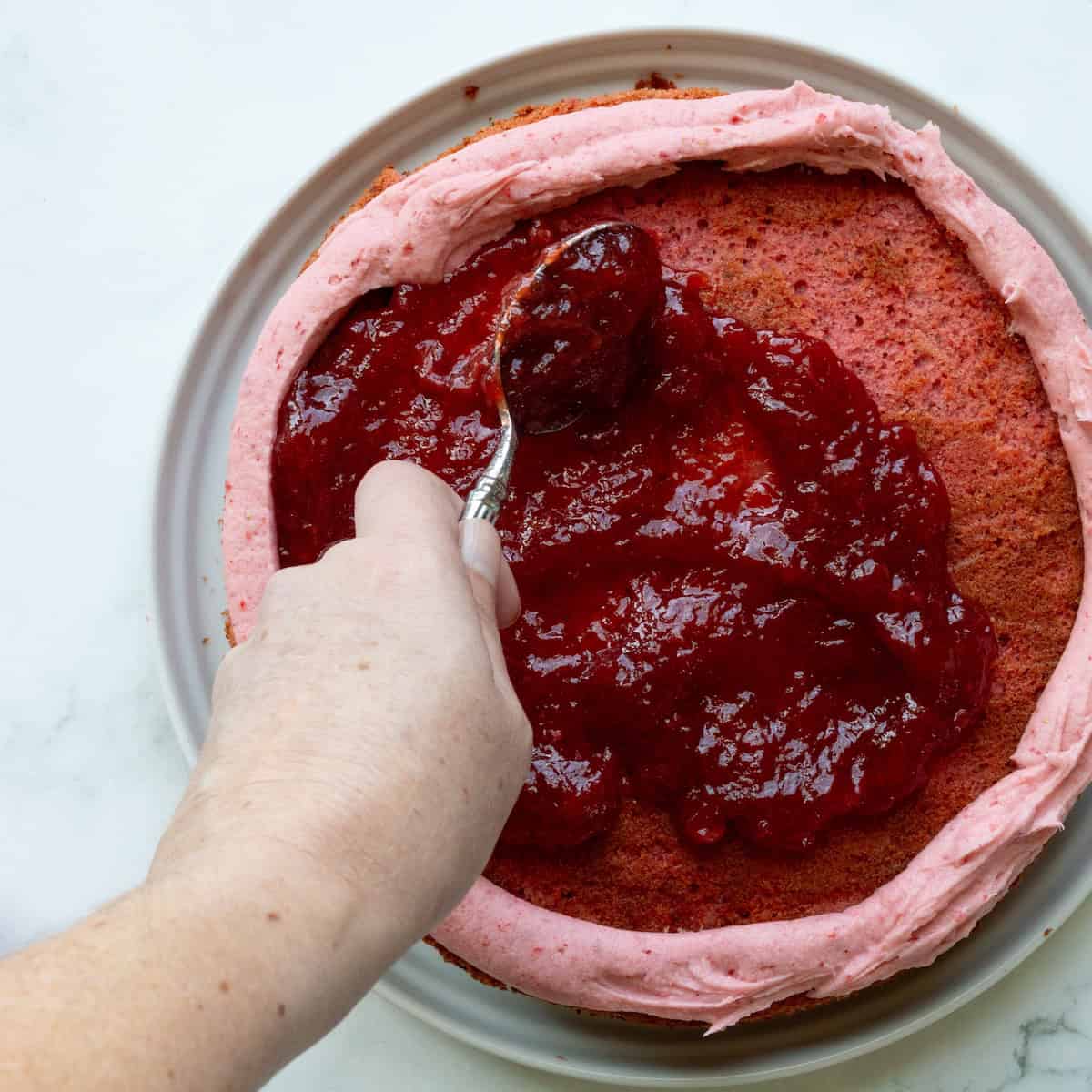 Spooning strawberry preserves onto a strawberry cake with a piped rim of strawberry buttercream.