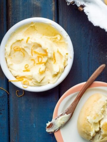 A white shallow bowl filled with honey butter with orange zest and a small bread plate with a roll and wooden spreader on the side.