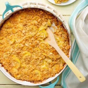 A round aqua baking pan of Cracker Barrel squash casserole with Ritz cracker topping and a wooden handled spoon scooped in.