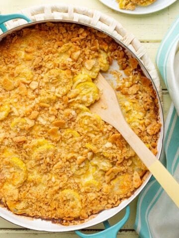 A round aqua baking pan of Cracker Barrel squash casserole with Ritz cracker topping and a wooden handled spoon scooped in.