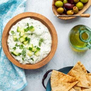 A wooden bowl of tzatziki dip with a bowl of pita wedges, a pitcher of olive oil, and olives on the side.