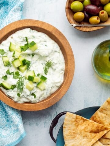 A wooden bowl of tzatziki dip with a bowl of pita wedges, a pitcher of olive oil, and olives on the side.