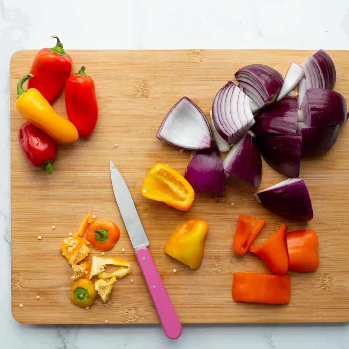 Cutting red onion and mini peppers into bite-size pieces on a wooden cutting board with a pink handled paring knife.