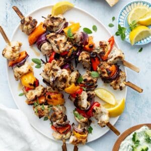Marinated Greek chicken kebabs on a white oval platter with homemade tzatziki and lemon wedges on the side.