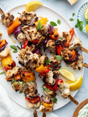Marinated Greek chicken kebabs on a white oval platter with homemade tzatziki and lemon wedges on the side.