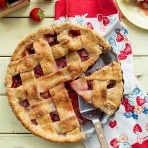 Lattice topped strawberry rhubarb pie with a slice on a plate and another slice being taken out.
