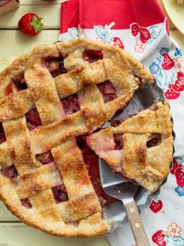 Lattice topped strawberry rhubarb pie with a slice on a plate and another slice being taken out.