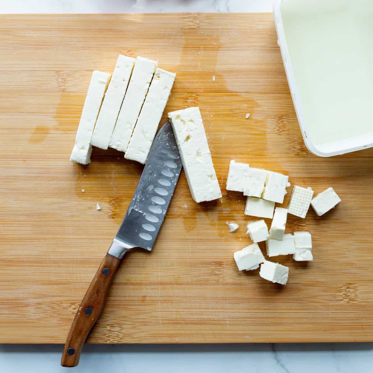 Cubing a block of fresh feta on a wooden cutting board.