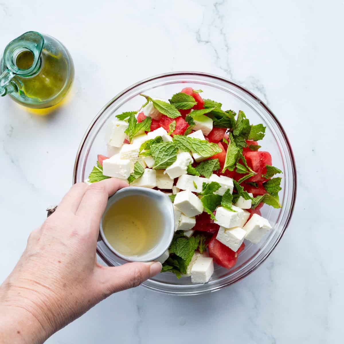 Pouring honey vinaigrette over cubed feta, fresh mint, and watermelon in a glass bowl.
