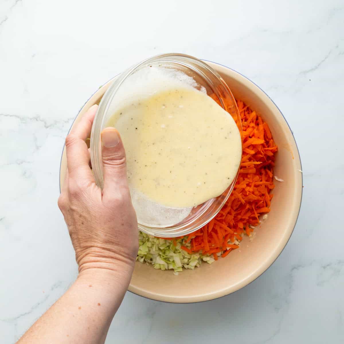 Pouring coleslaw dressing over carrots and cabbage in a large bowl.