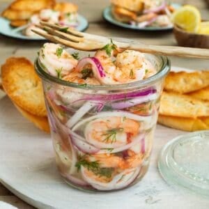 A glass jar of pickled shrimp with a wooden fork resting on top.