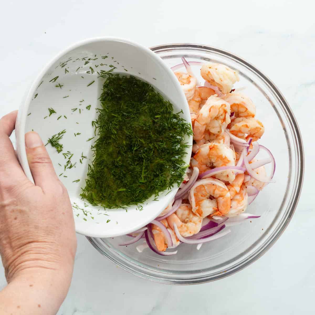 Pouring dill marinade over cooked shrimp and sliced red onion in a glass bowl.