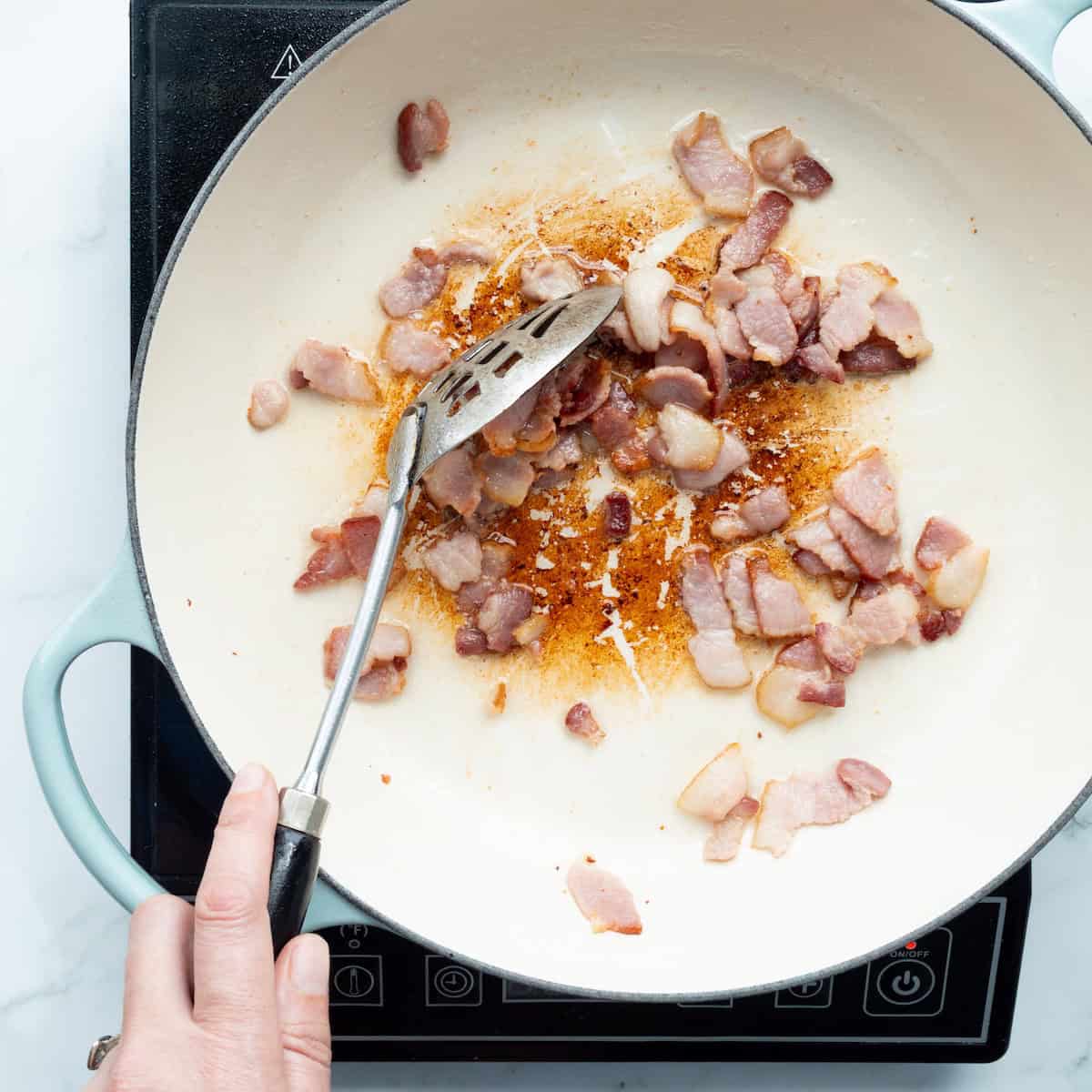 Cooking bacon pieces until crisp and stirring with a black handled slotted spoon.