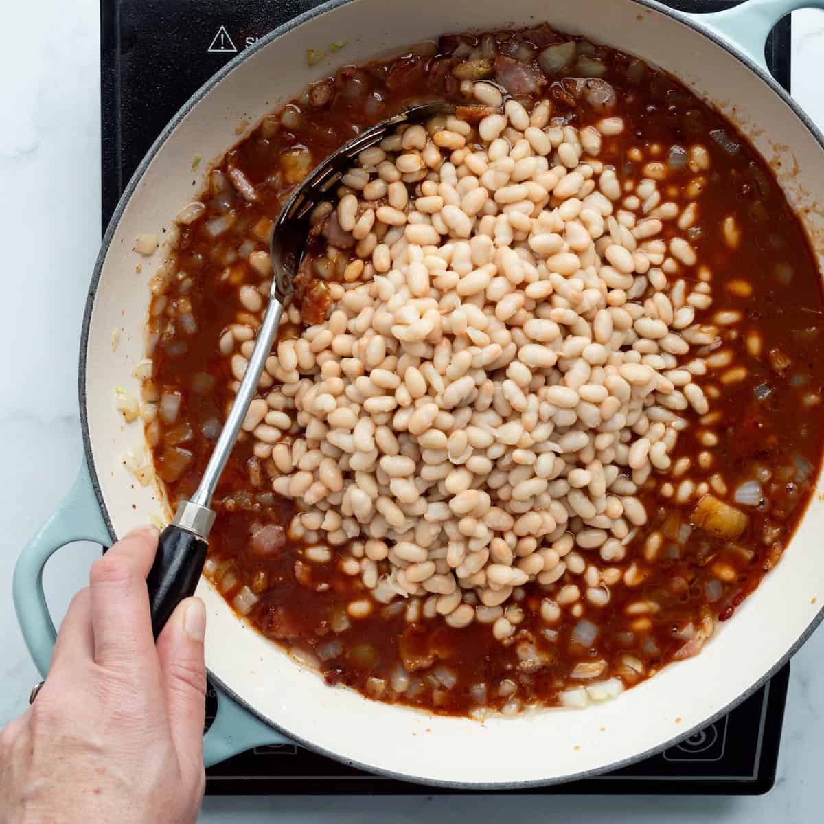 Stirring canned navy beans into a barbecue and bacon sauce in an aqua handled braiser.