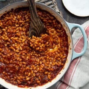 Barbecue baked beans in an aqua handled enamel braiser with a wooden spoon and red striped khaki napkin on the side.
