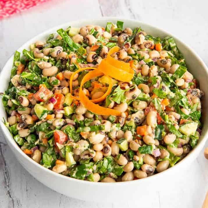 Black-eyed pea salad mixed with chopped collard greens and diced vegetables is served in a white bowl garnished with curled carrot ribbons.