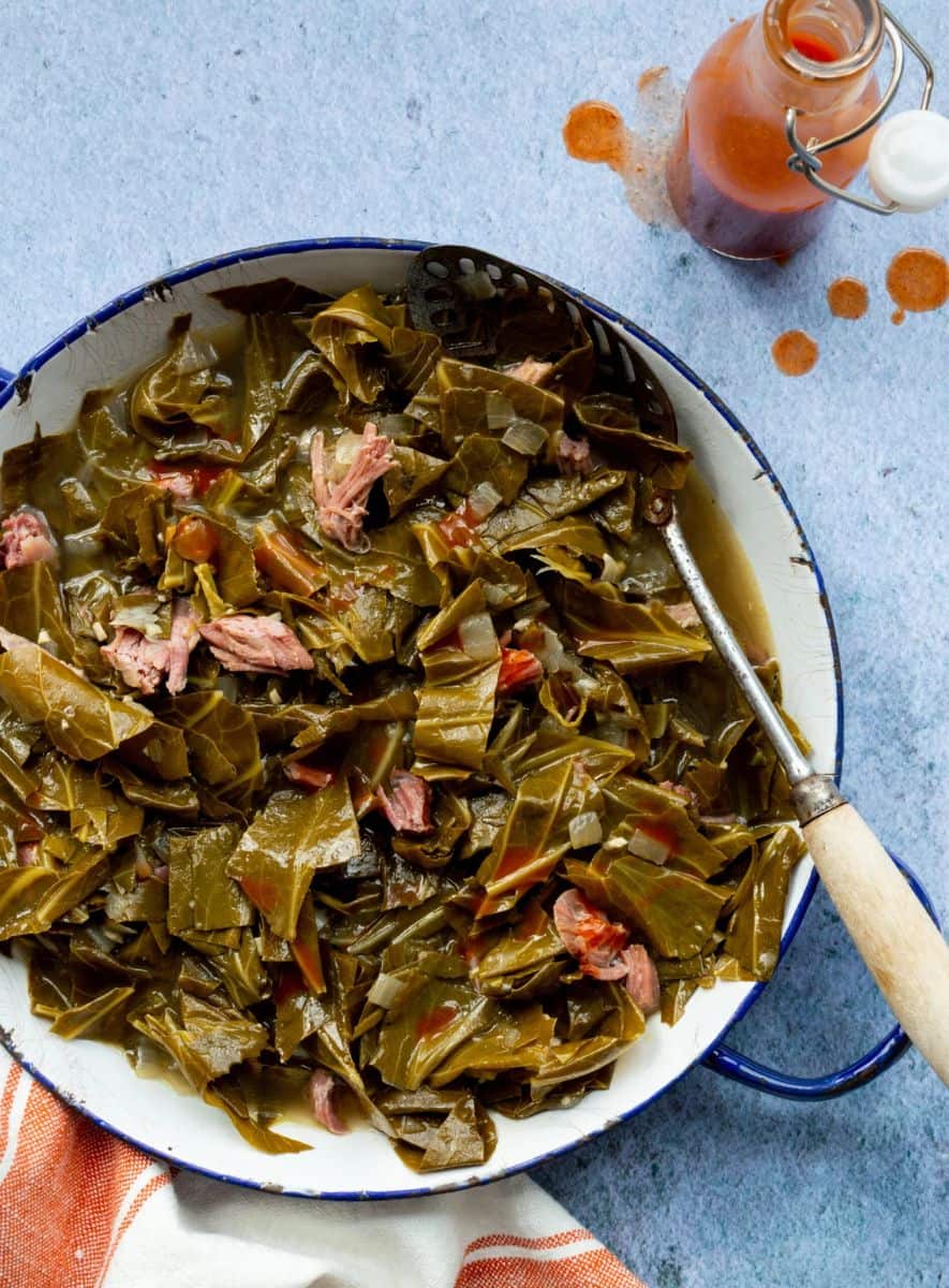Braised collard greens with tender pieces of smoked meat in a white enamel serving pot with a slotted spoon.