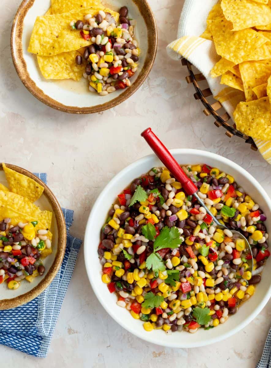 A white bowl of colorful Carolina caviar featuring corn, beans, and chopped peppers is served alongside a basket and small bowls filled with tortilla chips.