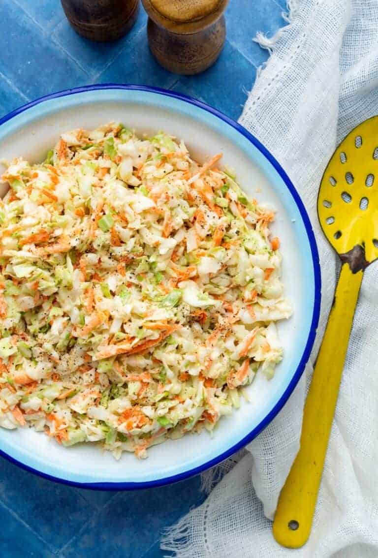 Creamy coleslaw in a white bowl, topped with black pepper, with a yellow serving spoon on the side.