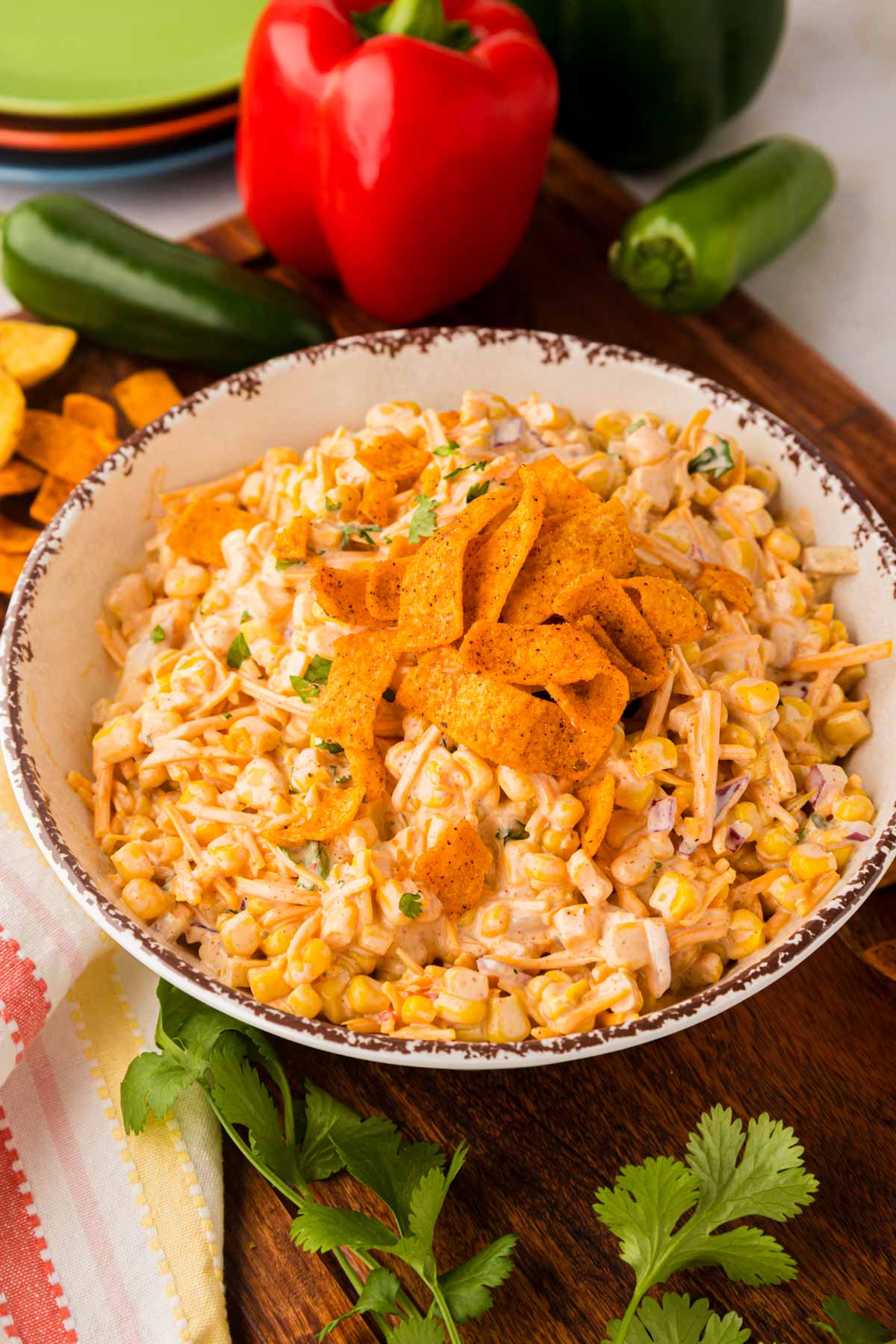 A white bowl filled with creamy frito corn salad topped with seasoned corn chips, set on a wooden surface with peppers and cilantro.
