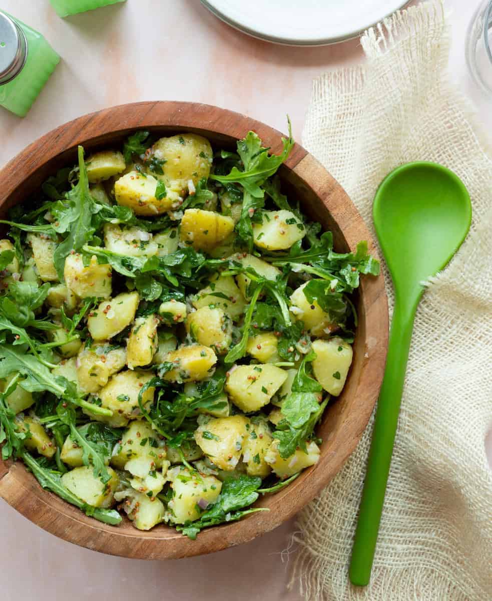 Potato salad with arugula and a mustard vinaigrette in a wooden bowl, served with a green spoon on the side.