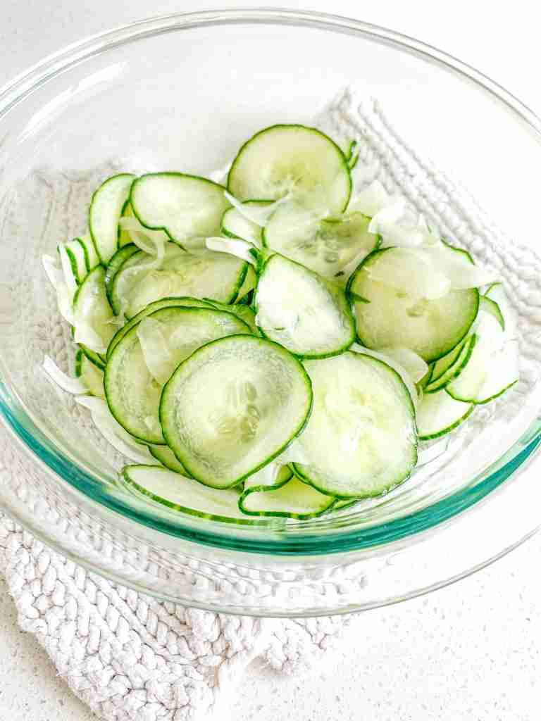 Large bowl filled with sliced cucumbers, placed on a white surface.