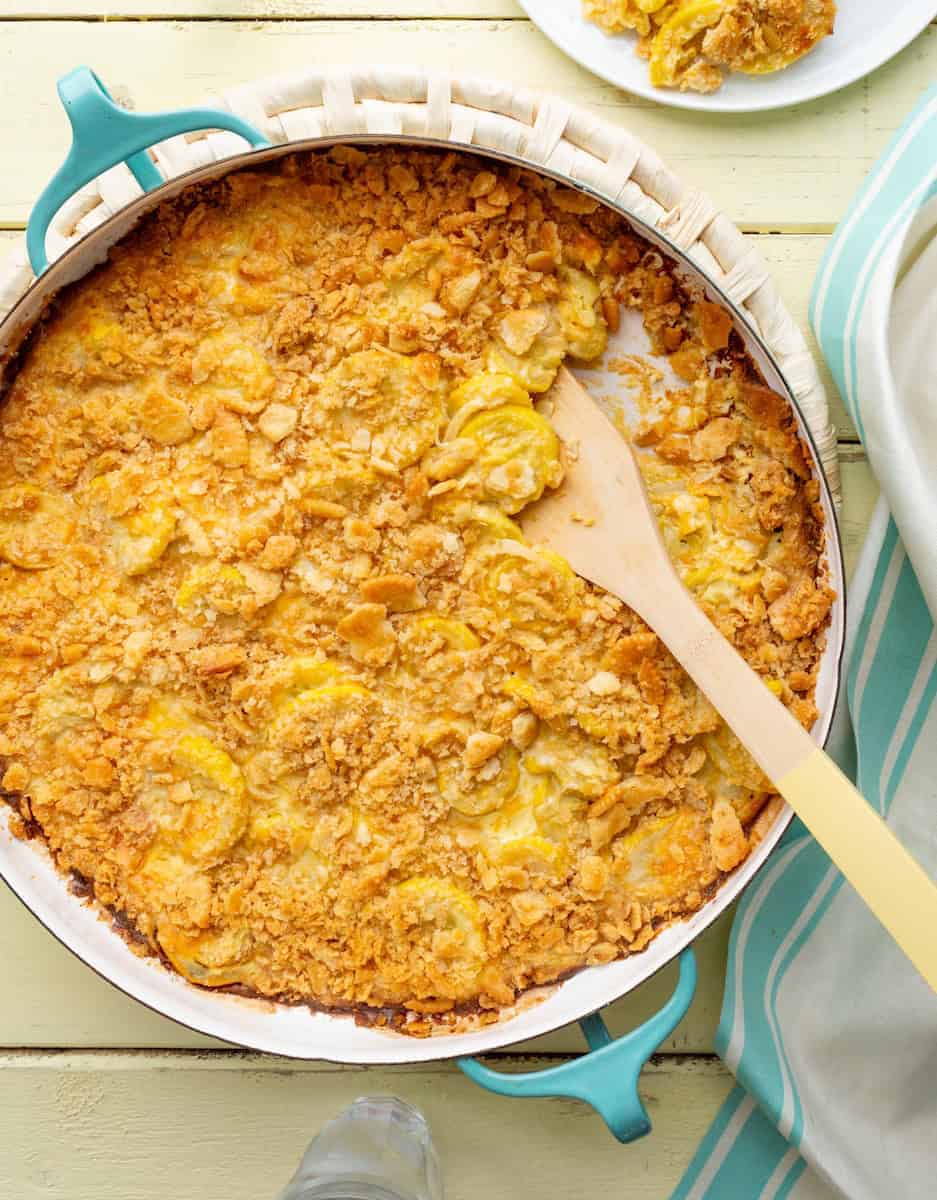 Golden squash casserole with a crispy cracker topping in a baking dish, served with a wooden spoon.